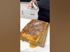 a person pours syrup over a cake on a cutting board in front of white cabinets