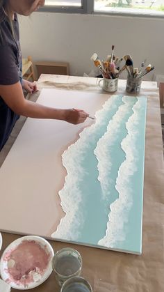 a woman is painting waves on a large piece of paper with watercolors in front of her