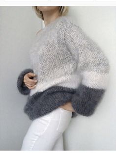 a woman wearing white pants and a sweater with grey stripes on the sleeves is leaning against a wall