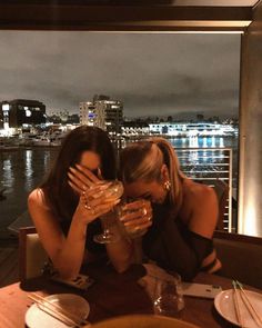 two women sitting at a table with wine glasses in front of their faces, looking out over the water