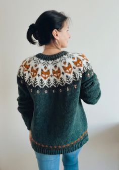 a woman standing in front of a white wall wearing a green sweater and blue jeans