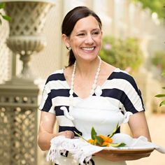 a woman holding a plate with flowers on it in front of a fountain and greenery