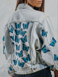 a woman wearing a jean jacket with blue butterflies on the front and back, standing in front of a white wall