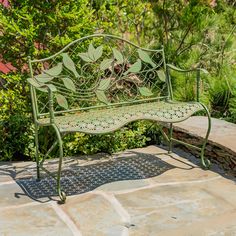 a green metal bench sitting on top of a stone walkway next to trees and bushes