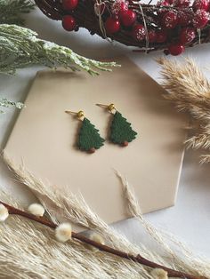 the earrings are made out of felt and have small trees on them, sitting next to some dried grass