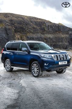 a blue toyota land cruiser parked in front of a rocky mountain side with the sky and clouds above it