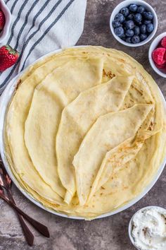 a plate filled with crepes next to bowls of strawberries and blueberries