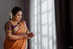 a woman standing in front of a window looking at her cell phone while wearing an orange and purple saree