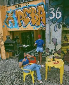 two women sitting in yellow chairs near graffiti covered walls and tables with drinks on them