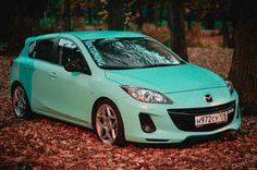 a blue mazda car parked in the woods with leaves on the ground and trees around it