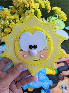 a person holding up a yellow and white sun shaped brooch with flowers in the background