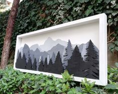 a white frame with trees and mountains cut out of it in front of some bushes