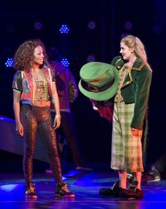 two girls in costumes on stage with one holding a green hat and the other looking at her