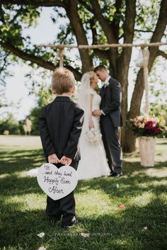 a little boy standing next to a woman in a wedding dress holding a heart shaped sign