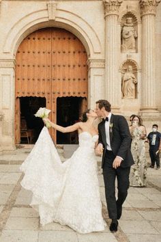 a bride and groom kissing in front of an ornate building with people standing around them