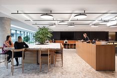 three people sitting at a table in an office setting with large windows and wooden desks