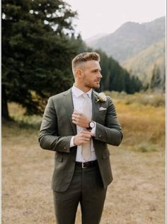 a man wearing a suit and tie standing in the middle of a field with mountains behind him