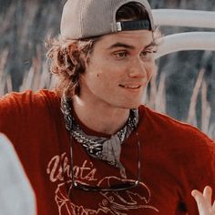 a young man wearing a baseball cap and red shirt talking to someone in the background