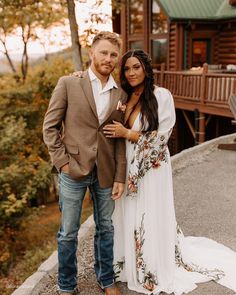 a man and woman standing next to each other in front of a log cabin at sunset