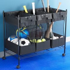 a black rack with four bins holding sports balls and tennis equipment on wheels in front of a blue wall