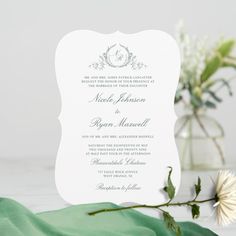 a white wedding card on top of a green cloth next to a vase with flowers