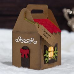 a small cardboard house with a red roof on top of snow covered ground next to a silver teapot