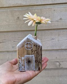 a hand holding a small wooden house with a flower in it's vase on the outside