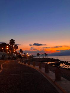 the sun is setting over the ocean with palm trees in the foreground and people walking on the sidewalk