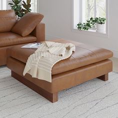 a brown leather ottoman with a blanket on it in front of a couch and potted plant