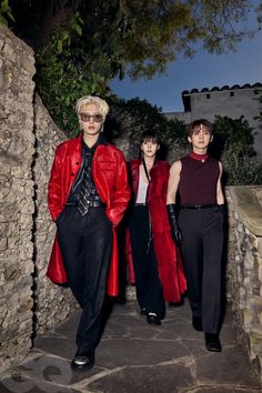 three people standing next to each other in front of a stone wall wearing red coats and black pants