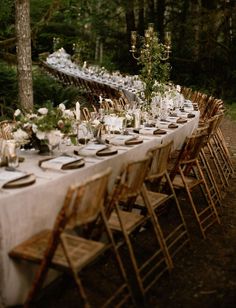 a long table is set up with white linens and gold place settings for dinner in the woods