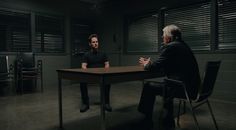 two men sitting at a table talking to each other in a dark room with shuttered windows