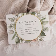 a white and gold wedding card with greenery on it, sitting on top of a blanket