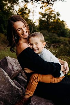 a woman holding a boy in her arms and smiling at the camera while sitting on some rocks