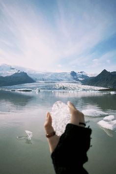 a person holding up a piece of ice in front of some water with mountains in the background