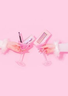 two hands holding wine glasses filled with pink liquid and some writing on the paper next to them