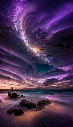 the sky is filled with purple clouds and stars as people stand on rocks in the water