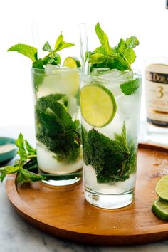 two glasses filled with mojito and limeade on a wooden tray next to a bottle