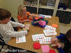 three children sitting at a table working on matching shapes and numbers with paper cutouts