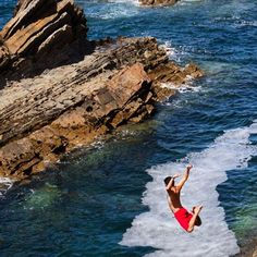 a man in red swimsuit jumping off rocks into the ocean from a cliff above