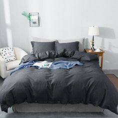 a bed with grey sheets and pillows in a white room next to a lamp on a table