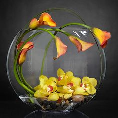 an arrangement of flowers in a glass bowl on a reflective surface with water and pebbles