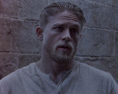 a man standing in front of a brick wall wearing a grey shirt and looking at the camera