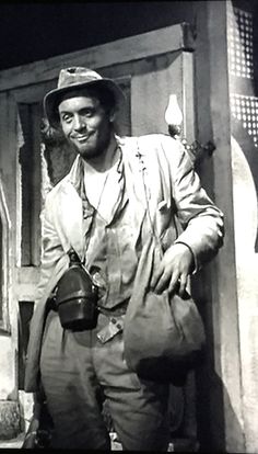 a man wearing a hat and holding a camera in front of a doorway with an open door