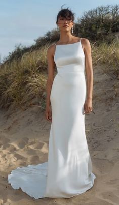 a woman standing in the sand wearing a white wedding dress with one shoulder cut out