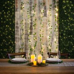 the table is set for dinner with candles and greenery on the wall behind it