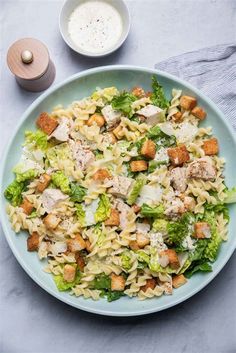 a pasta salad with chicken and broccoli on a blue plate next to a bowl of ranch dressing