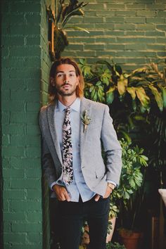 a man in a suit and tie leaning against a green wall with plants behind him