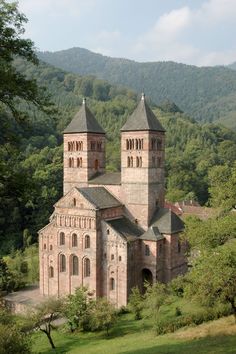 an old building with two towers in the middle of trees and hills behind it on a sunny day