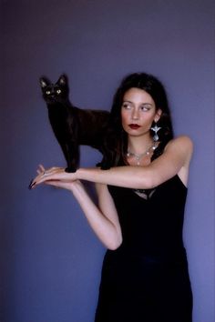 a woman holding a black cat on her arm in front of a purple wall with a blue background